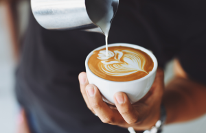 Ihminen kaataa vaahdotettua maitoa kahvikuppiin, ja pinnalle muodostuu kaunista latte art -kuviota. 