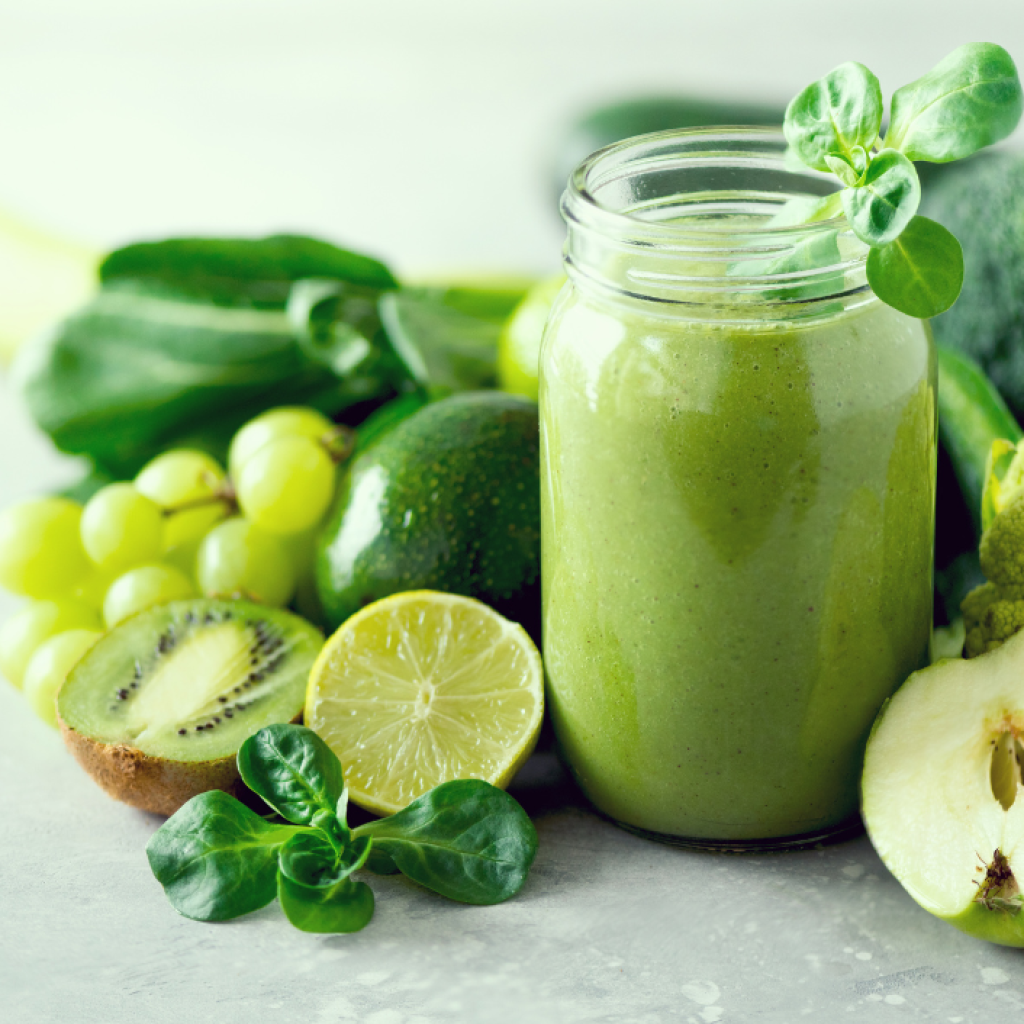 Vihersmoothie lasipurkissa, taustalla vihreitä hedelmiä ja vihanneksia kuten kiiviä, limeä, omenaa, avokadoa ja parsakaalia.