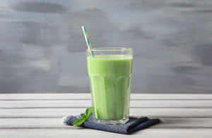 Korkea lasi vihersmoothieta, jossa vihreä raidallinen pilli ja koristeena mintunlehti. Lasin alla tummansininen kangasliina, taustalla harmaansininen maalattu seinä.