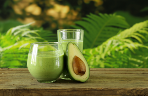 Vihreä avokadosmoothie lasipurkissa, jossa on vaaleanpunavalkoinen pilli. Taustalla näkyy avokadoja ja ananaspaloja vaaleansinisellä pöydällä.