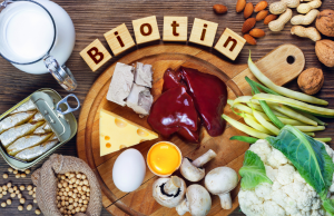 Pöydällä eri biotiinipitoisia ruoka-aineita, kuten maksa, kananmuna, juusto, maito, sardiinit, pavut, kaali, sienet ja pähkinät. Keskellä puiset palikat muodostavat sanan "Biotin".

