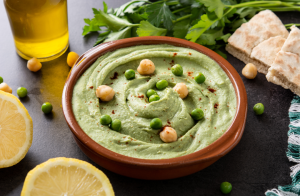 Hernehummus tarjoiltuna ruskeassa kulhossa, koristeltuna vihreillä herneillä ja kikherneillä, ympärillä yrttejä ja sitruunaviipaleita.