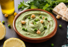 Hernehummus tarjoiltuna ruskeassa kulhossa, koristeltuna vihreillä herneillä ja kikherneillä, ympärillä yrttejä ja sitruunaviipaleita.