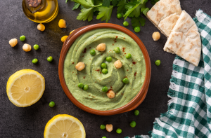 Ylhäältä kuvattu vihreä hernehummus kulhossa, koristeltuna herneillä ja kikherneillä, ympärillä pita-leipää, sitruunaa ja yrttejä.