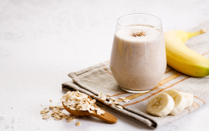 Banaani-kaurasmoothie tarjoiltuna lasissa kaurahiutaleiden ja tuoreen banaanin kera vaalealla keittiöalustalla.