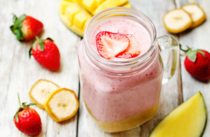 Vaaleanpunainen smoothie lasipurkissa, päällä mansikkaviipaleita. Taustalla banaania, mansikoita ja mangoa.