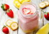 Vaaleanpunainen smoothie lasipurkissa, päällä mansikkaviipaleita. Taustalla banaania, mansikoita ja mangoa.