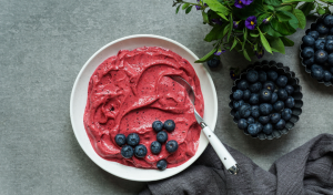 Valkoisella lautasella on paksua, marjaisaa nice cream -jäätelöä, jonka päällä on tuoreita mustikoita. Vieressä näkyy pieniä metallikulhoja täynnä mustikoita ja vihreitä yrttejä.