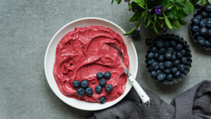 Valkoisella lautasella on paksua, marjaisaa nice cream -jäätelöä, jonka päällä on tuoreita mustikoita. Vieressä näkyy pieniä metallikulhoja täynnä mustikoita ja vihreitä yrttejä.