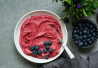 Valkoisella lautasella on paksua, marjaisaa nice cream -jäätelöä, jonka päällä on tuoreita mustikoita. Vieressä näkyy pieniä metallikulhoja täynnä mustikoita ja vihreitä yrttejä.