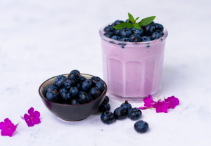 Lasi vaaleanviolettia mustikkavaahtoa, jonka päällä tuoreita mustikoita ja mintunlehti. Vieressä pieni kulho mustikoita ja koristeena pinkkejä kukkia.

