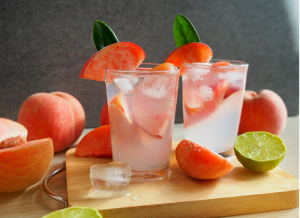 Kaksi lasillista vaaleanpunaista peach mocktailia jääpaloilla ja persikkakoristeilla puualustalla, ympärillä persikoita ja limenpuolikkaita.