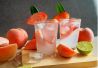 Kaksi lasillista vaaleanpunaista peach mocktailia jääpaloilla ja persikkakoristeilla puualustalla, ympärillä persikoita ja limenpuolikkaita.