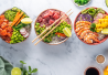 Kolme värikästä sushibowlia limelohkoilla koristeltuna; kulhoissa lohta, tonnikalaa, katkarapuja, mangoa, avokadoa, edamamea ja kasviksia.