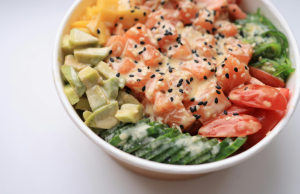 Lähikuva lohipohjaisesta sushibowl-annoksesta, jossa on avokadoa, mangoa, merilevää, tomaattia, kurkkua ja seesamkastiketta.