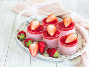 Lähiotoksessa viisi pientä jälkiruokakuppia, joissa vaaleanpunainen mousse ja päällä mansikka-vadelmakastiketta sekä puolikas mansikka koristeena. Ympärillä tuoreita mansikoita ja kaksi lusikkaa vaalealla puisella alustalla.