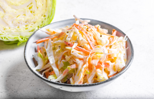 Raikas coleslaw-salaatti vaaleassa kulhossa valkoista taustaa vasten, kaalinpuolikas taustalla.