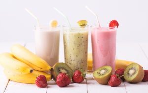 Kolme smoothielasia banaanilla, kiivillä ja mansikalla koristeltuna, edessä tuoreita hedelmiä.