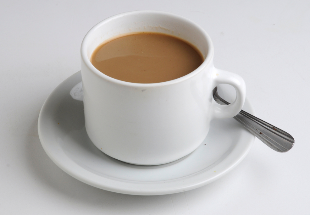 Valkoinen kahvikuppi, jossa maitokahvia ja lusikka lautasella.
