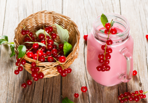 Punertava smoothie lasipurkissa, jonka päällä punaherukoita ja mintunlehtiä. Vieressä pieni kori täynnä tuoreita punaherukoita. Taustalla vanha puupöytä.