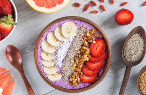 Puukulhossa tarjottu violetin sävyinen smoothiebowl, koristeltuna banaanilla, mansikalla, granolalla ja siemenillä.