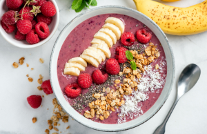 Banaania, vadelmia ja granolaa sisältävä smoothiebowl valkoisella pöydällä, vieressä banaaneja ja vadelmakulho.

