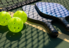 Kaksi pickleball-mailaa kentällä, joiden vieressä kolme pickleball-palloa. Peliverkko muodostaa varjon mailojen päälle.