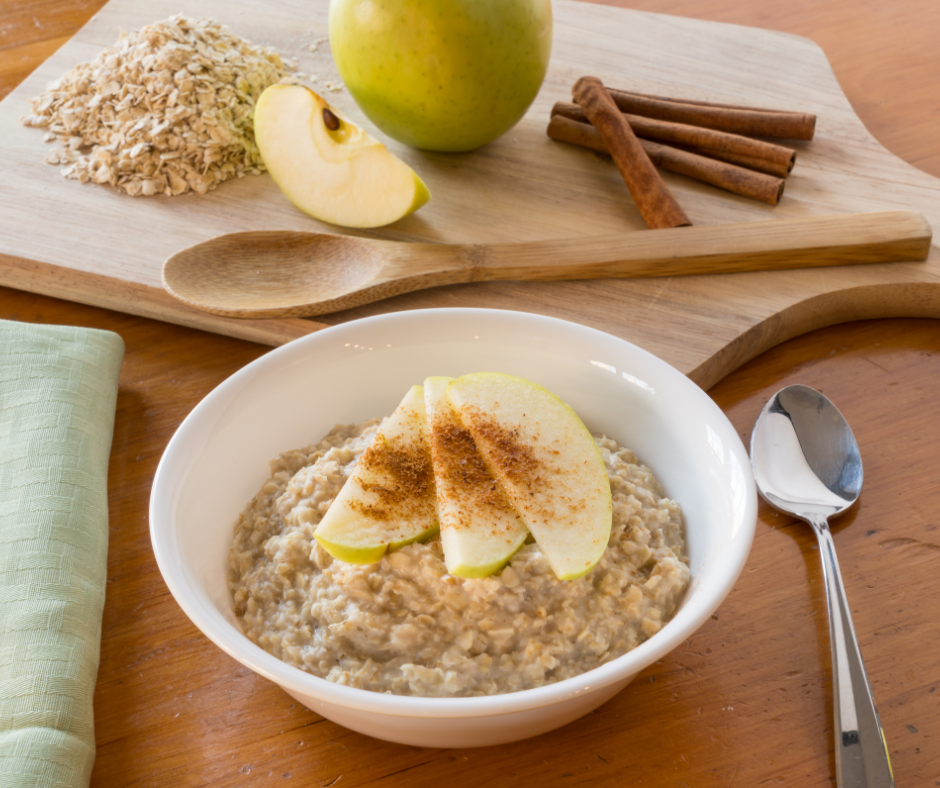 Kuvassa lautasellinen kaurapuuroa, jossa on päällä omenaviipaleita ja kanelia. Taustalla leikkuulaudan päällä vihreä omena, kanelitankoja ja kaurahiutaleita.