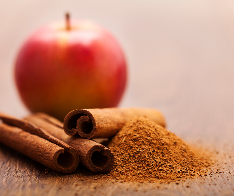 Kuvassa jauhettua kanelia ja kolme kanelitankoa pöydällä. Taustalla punainen omena.