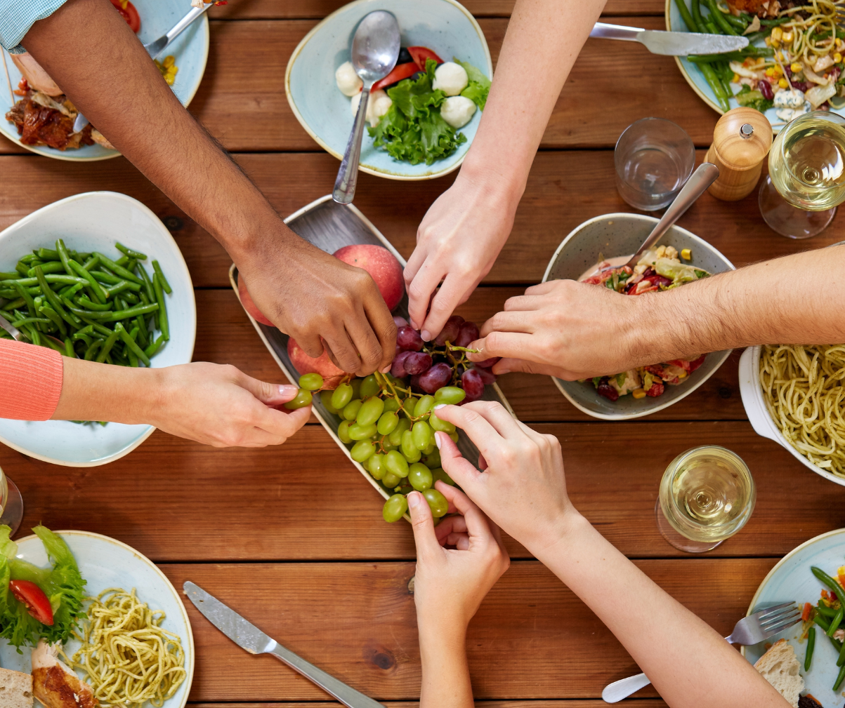 Kuva on otettu ylhäältäpäin ja siinä istuu joukko ruokailijoita ruokapöydän ääressä. He kaikki ovat ottamassa samaan aikaan rypäleitä.