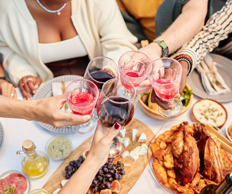 Kuvassa ruokapöydän ääressä olevat henkilöt kilistelevät viinilaseilla. Ruokapöydällä näkyy myös herkullisia ruokia kuten juustoja ja uunissa paistettua lihaa.