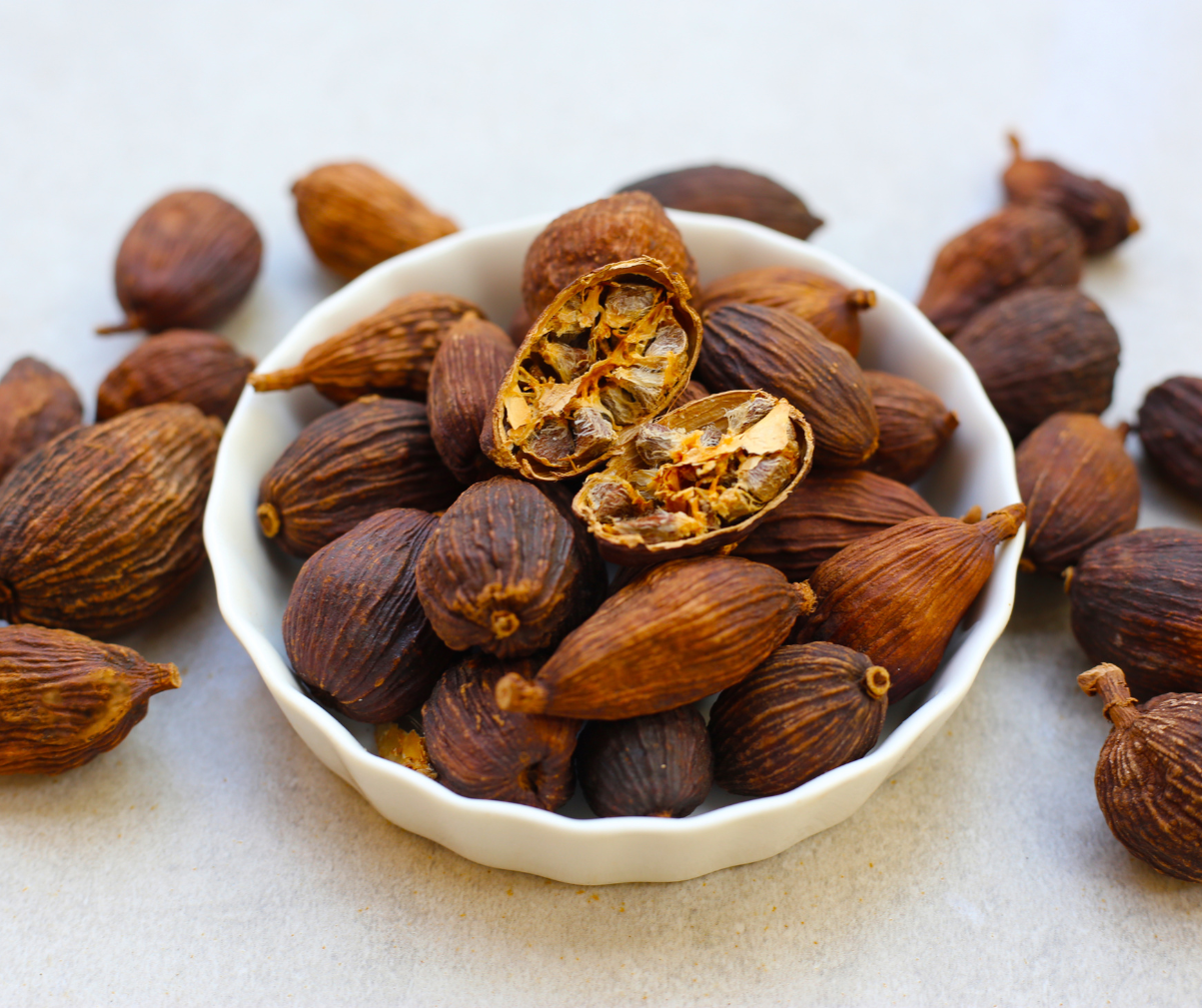 Kuvassa ruskeaa kardemummaa valkoisessa pienessä kulhossa ja sen ympärillä.