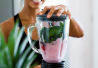 Kuvassa nainen valmistaa smoothieta ioisena. Smoothieblenderi on kuvan etualalla ja nainen taustalla