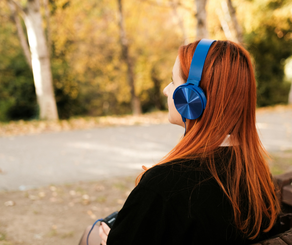 Kuvassa henkilö kuuntelee äänikirjaa- tai podcastia kävelyllä. Taustalla syksyinen luonto. Naisella on punaiset hiukset ja siniset kuulokkeet.