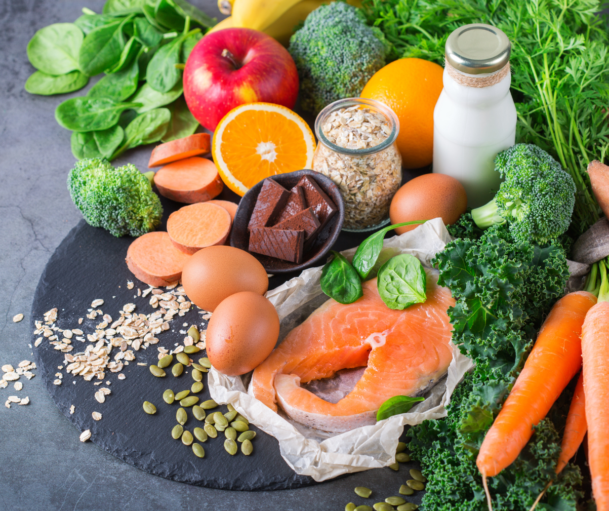 Kuvassa erilaisia vitamiineja sisältäviä raaka-aineita kuten lohta parsakaalta, kananmunia, sitruksia, omenoita, pinaattia ja bataattia