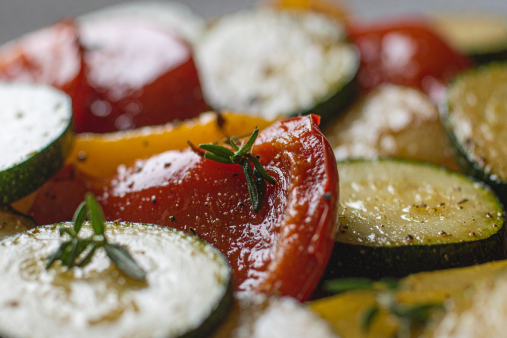 Lähikuva uunissa paahdetuista kasviksista: paprikan, kesäkurpitsan ja tomaatin viipaleita maustettuna yrteillä ja öljyllä.