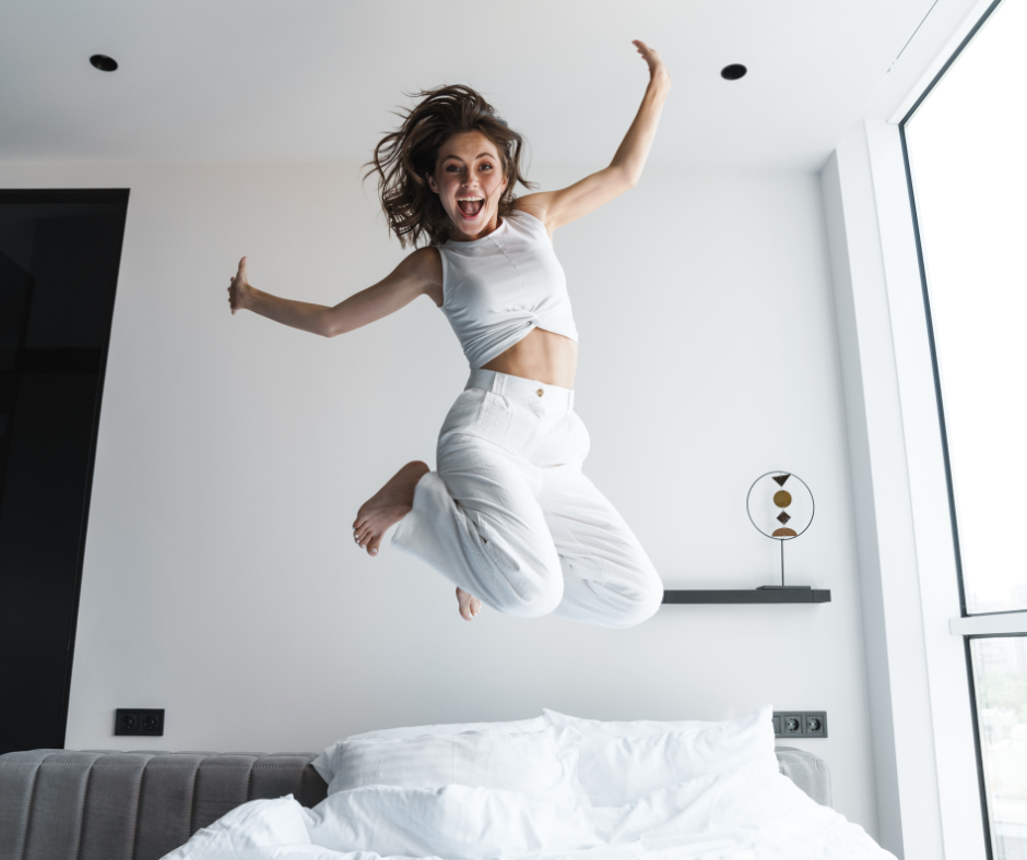 Iloinen nainen hyppää sängyllä vaaleassa makuuhuoneessa, pukeutuneena valkoisiin vaatteisiin ja kädet ilmassa.