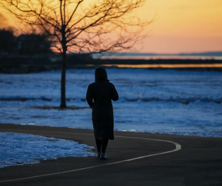 Ihminen kävelee talvisessa maisemassa iltahämärässä, luminen maa ja auringonlasku taustalla.