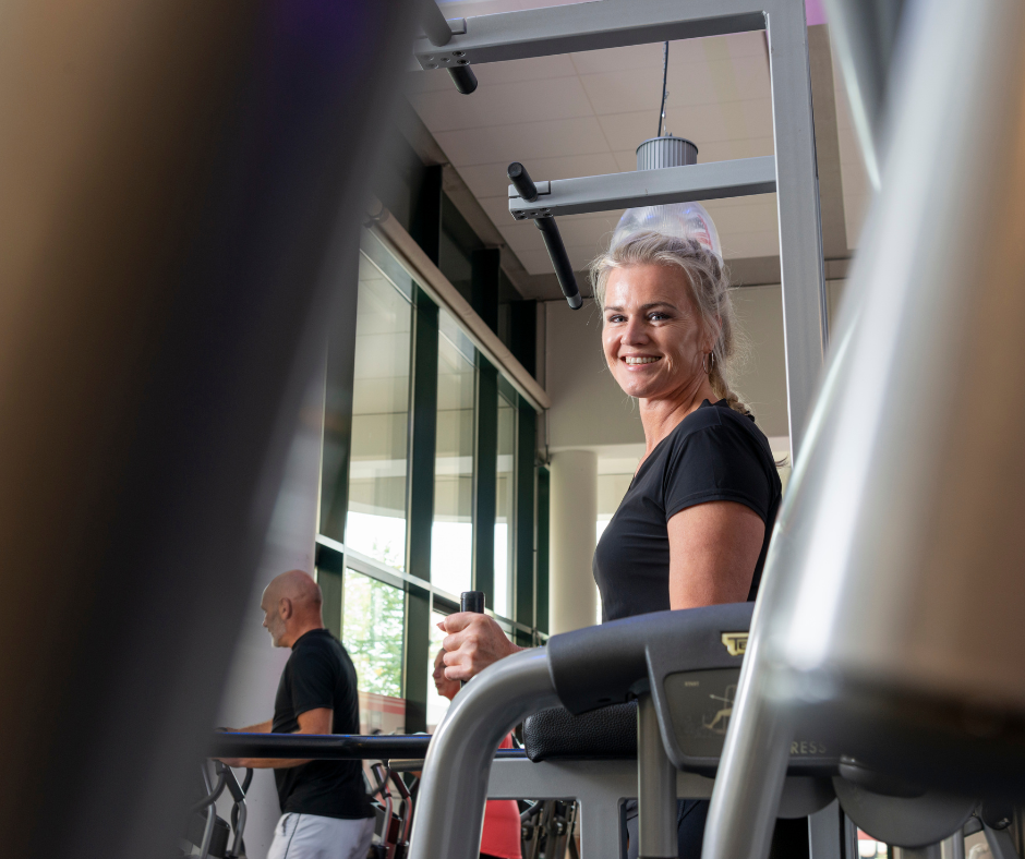 Nainen hymyilee kuntosalilla. Kahden laitteen välistä kuvattuna.