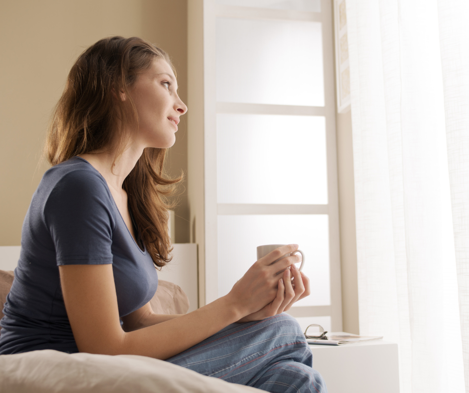 Nainen istuu sängyllä kahvikuppikädessä ja katsoo ulos.