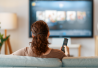 Nainen istuu sohvalla ja katsoo televisiota kaukosäädin kädessä.