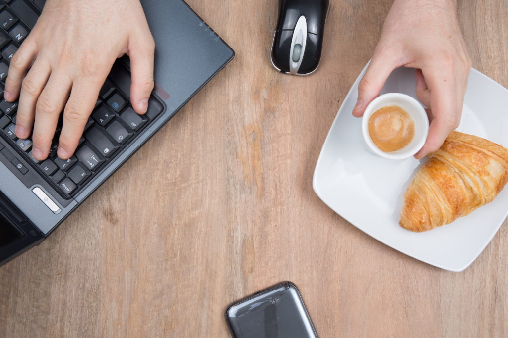 Henkilö tekee töitä tietokoneella ja syö samaan aikaan croissanttia ja juo espressoa.