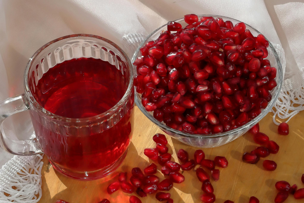Läpinäkyvä muki täynnä punaista granaattiomenamehua sekä kulhollinen granaattiomenan siemeniä valoisalla pöydällä.