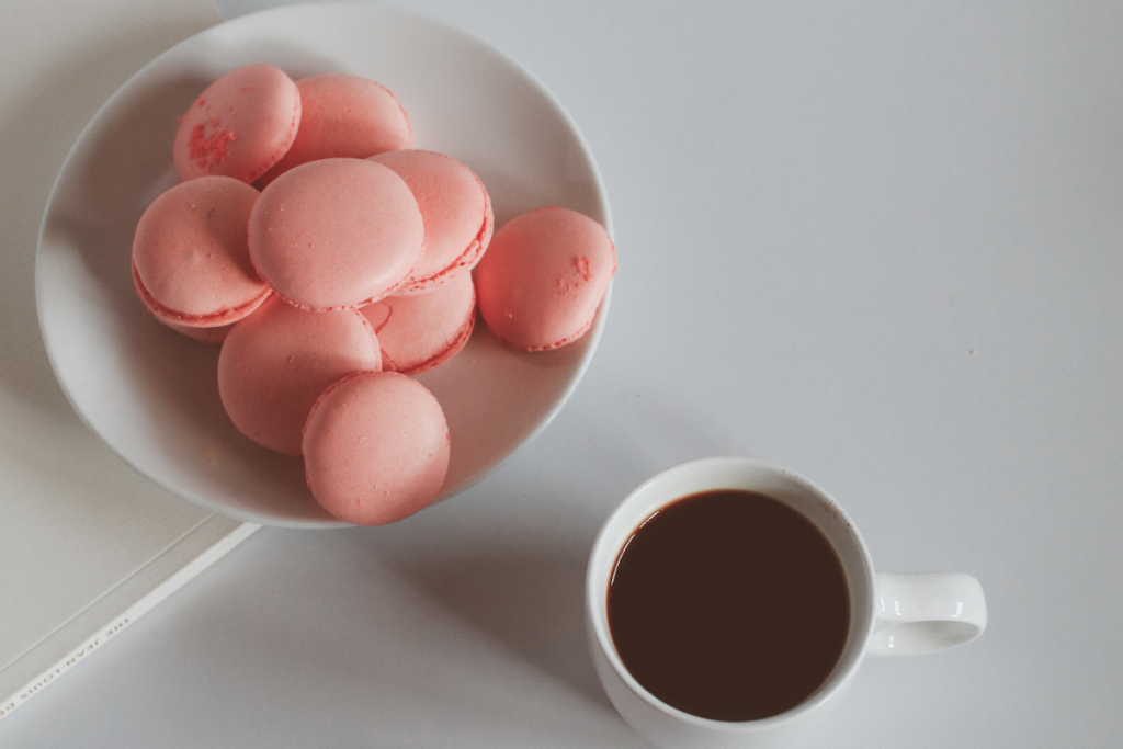 Kulhossa vaaleanpunaisia macarons-leivoksia ja vieressä kuppi kahvia.