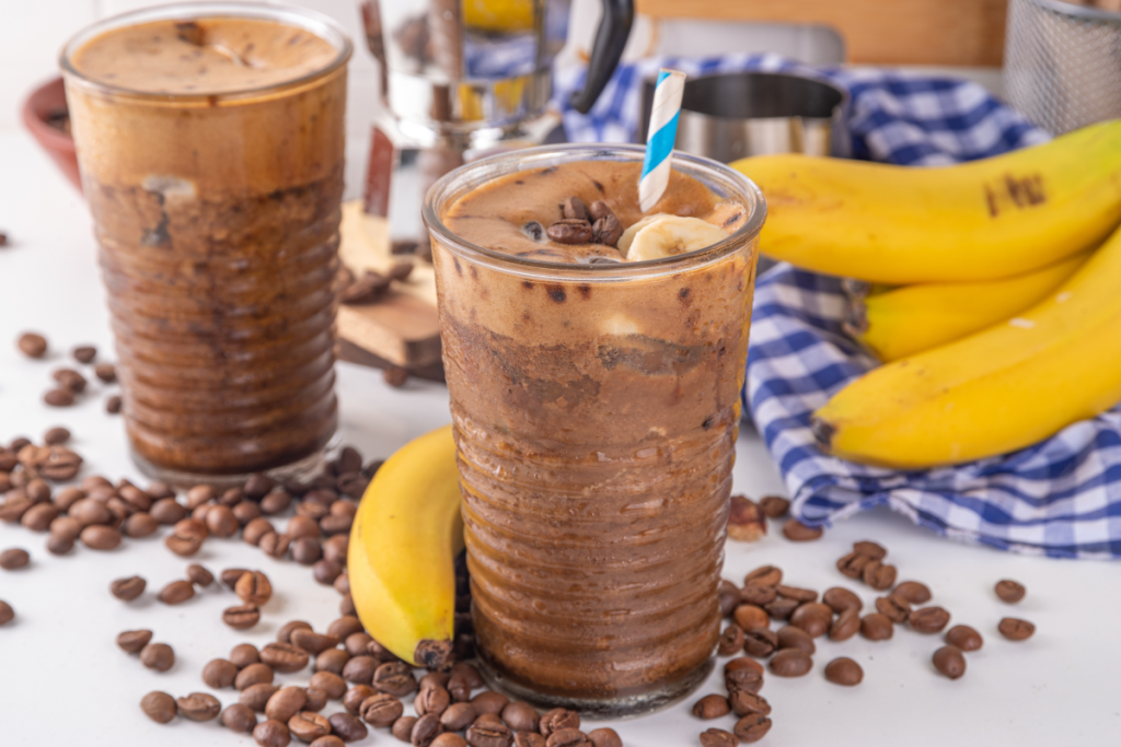 Banaanilatte-juomia. Vieressä kahvipapuja ja kokonaisia banaaneja.