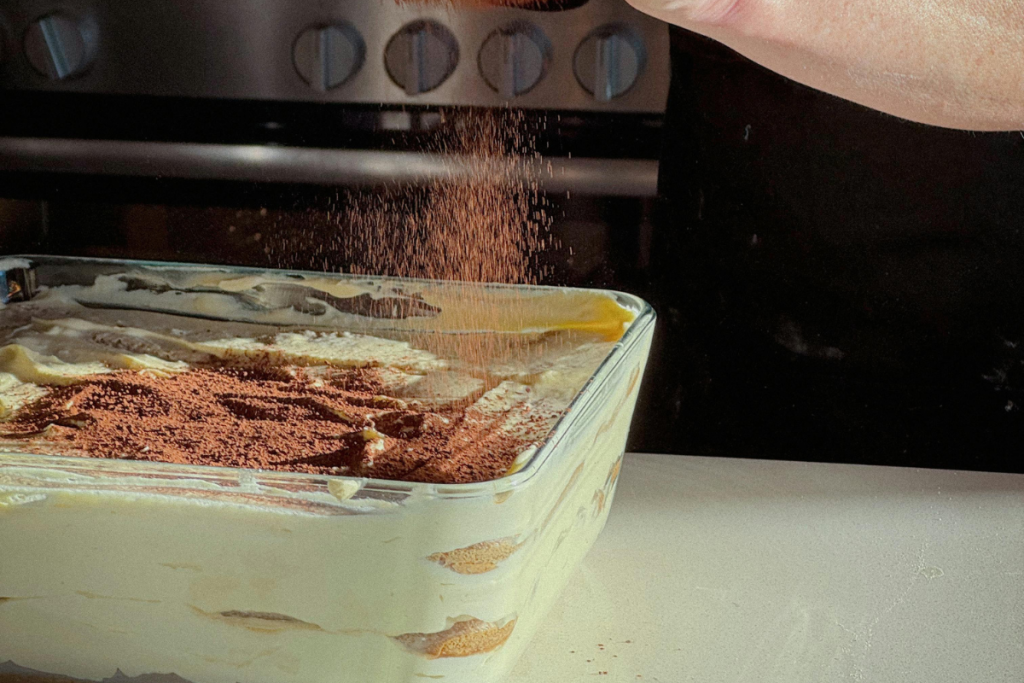 Tiramisu vuoassa, päälle ripotellaan kaakaojauhetta.