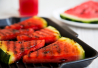 Grillattuja vesimeloneja lautasella. Taustalla mehua ja vesimelonin pala.